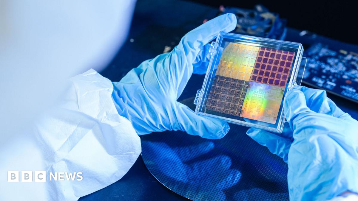 An engineer in a cleanroom, wearing blue gloves and white overalls, inspects a sheet of semiconductors