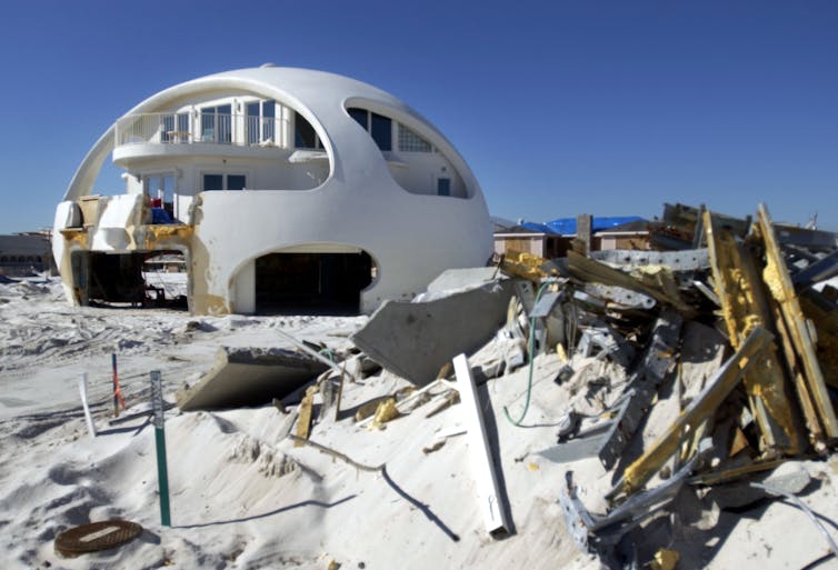 A house with a concrete dome and cutouts for windows sits on the beach, mostly in good shape, with storm debris around it.
