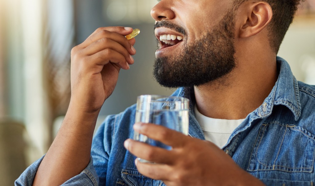 Man holding a vitamin capsule to his mouth with a glass of water in his other hand.