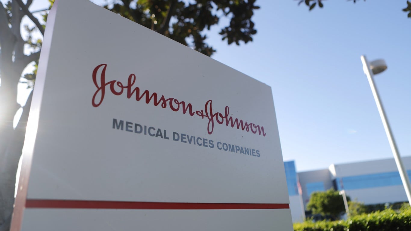 A close-up, low-angle shot of a white rectangular corporate sign for Johnson & Johnson. The company's name is written in a distinctive red script, with 'MEDICAL DEVICES COMPANIES' in a smaller, gray sans-serif font below. Sunlight creates a bright lens flare on the left side of the sign, with blurred green tree foliage and a clear blue sky in the background. A white light pole is visible on the right, and parts of a modern building with blue-tinted windows can be seen in the distance.