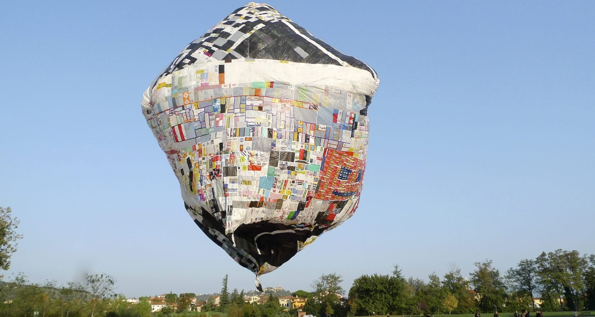a floating artwork by Tomás Saraceno featuring a patchwork object resembling an unusual air balloon over a square