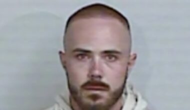 A mug shot of a man with a shaved head, light skin, and a short, dark beard, looking directly at the camera