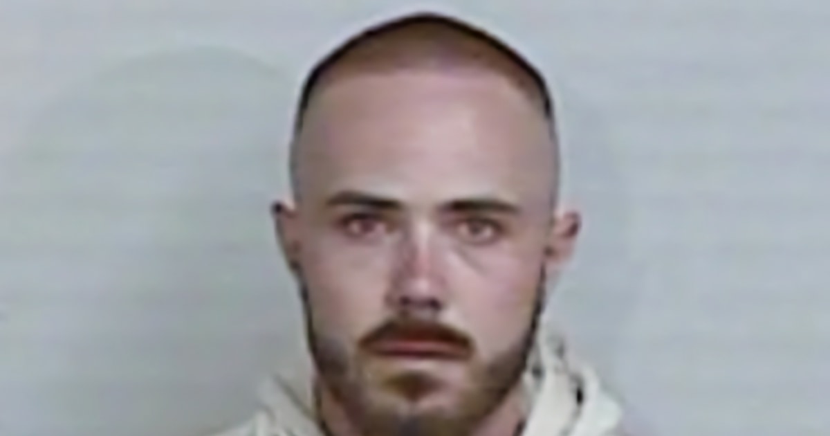 A mug shot of a man with a shaved head, light skin, and a short, dark beard, looking directly at the camera