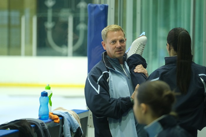 The Blame A man in a sports jacket helps a woman stretch her raised leg in an indoor ice rink or sports facility. Two water bottles and sports gear sit nearby.