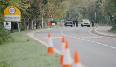 Road lined with traffic cones and a 30 km/h speed limit sign at the entrance to Whelford, with police vehicles and officers visible in the distance.
