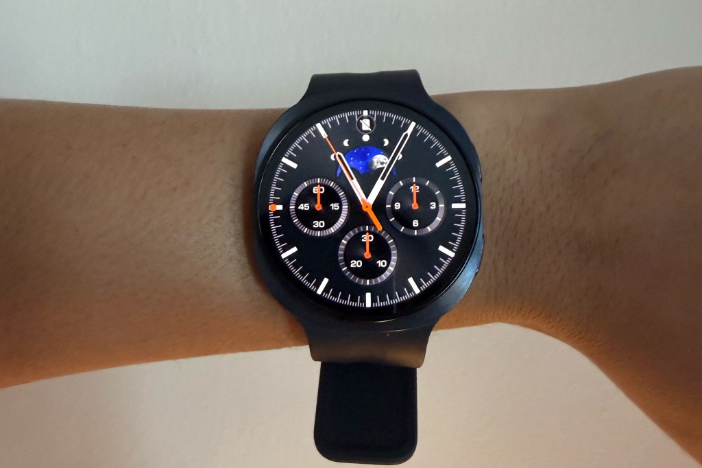 A black smartwatch with a complex watch face featuring three sub-dials and a moon phase indicator, worn on a person's wrist.