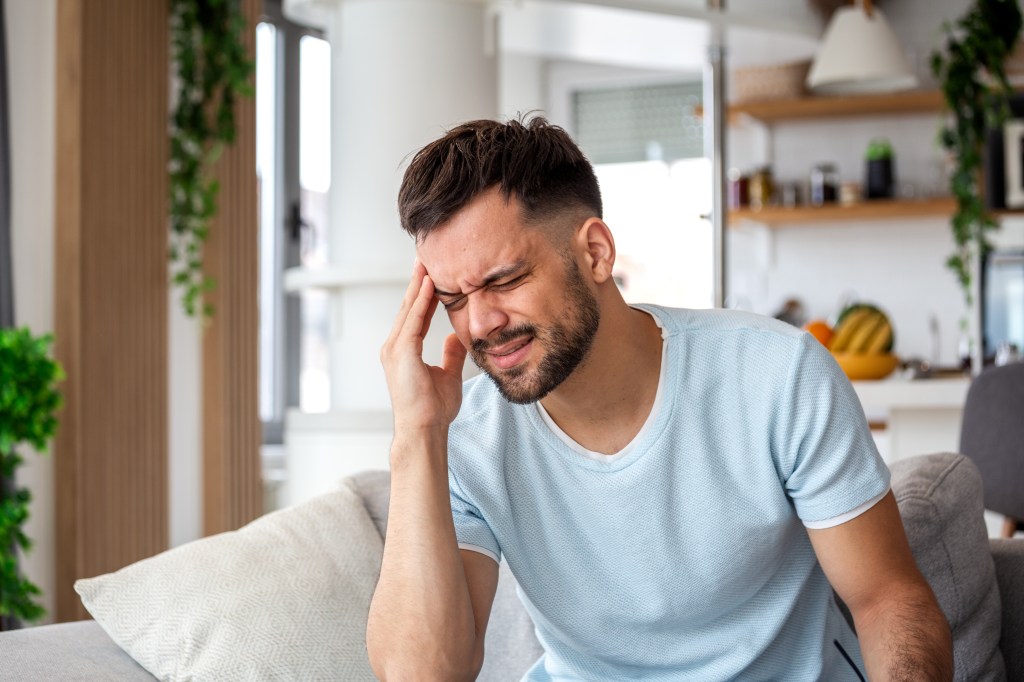 A young man with a beard sits on a couch, holding his head and grimacing in pain from a severe headache.