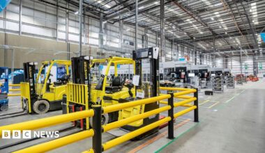 A large warehouse has shelving and other industrial fittings inside and some yellow and black forklift trucks are in a section surrounded by yellow railings.