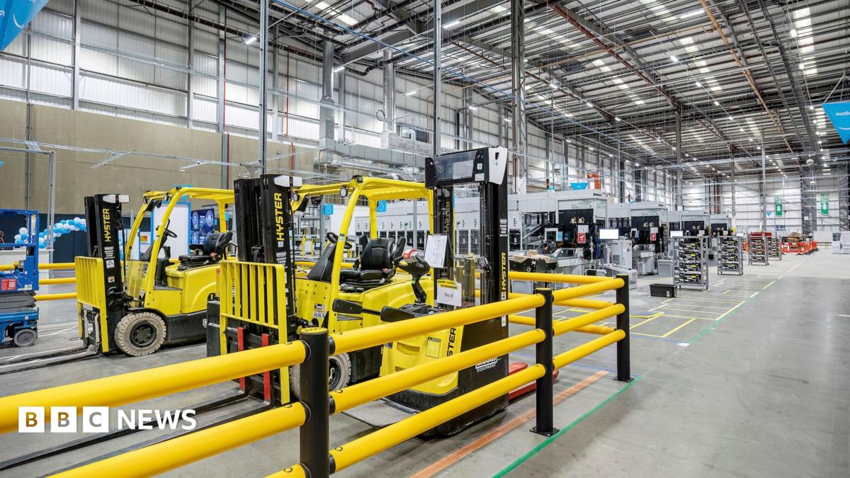 A large warehouse has shelving and other industrial fittings inside and some yellow and black forklift trucks are in a section surrounded by yellow railings.