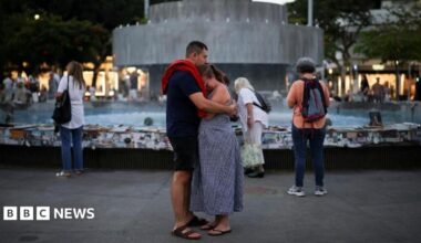 A man and woman embrace in front of the fountain at Dizengoff Square. The woman has her hand to her eyes as if she is crying.