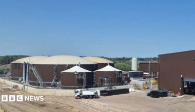 An industrial-looking site dominated by three large, circular, brown storage tanks with white domed tops, next to a large brown warehouse.