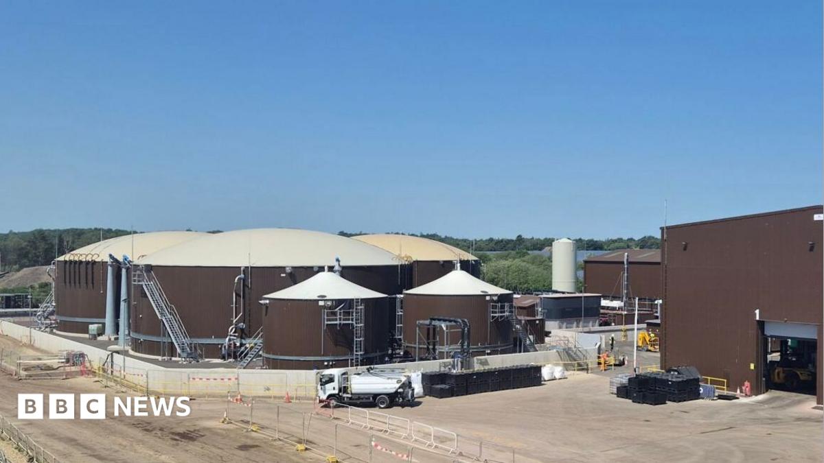 An industrial-looking site dominated by three large, circular, brown storage tanks with white domed tops, next to a large brown warehouse.