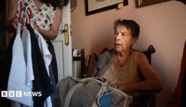 Maricarmen Abascal sits in a chair in her flat ahead of her eviction, in Madrid, Spain on 23 September. She has short, dark hair and is wearing a cream vest. A holdall bag is on her lap. A woman whose face is out of shot is holding up clothes on a hanger next to Abascal.