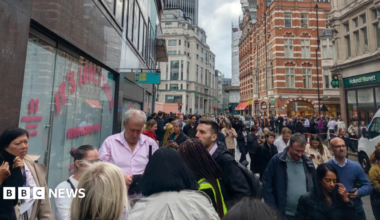 Crowds of people stand on a pavement and spill into the road on a street in the City of London, surrounded by office buildings and shops.