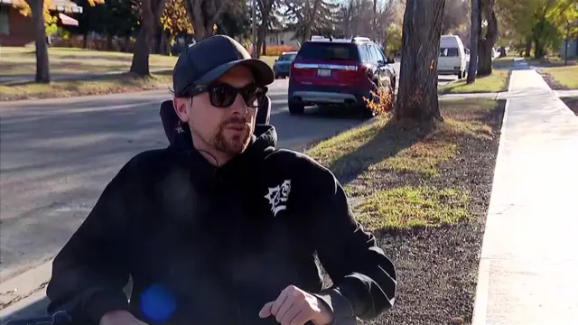 A man is sitting in a wheelchair, with his arms on the arm rest, on a pavement next to what appears to be a residential street with several trees and cars parked next to the curb. The man has a black goatee and is wearing a black cap with black tinted glasses and a black hoodie.