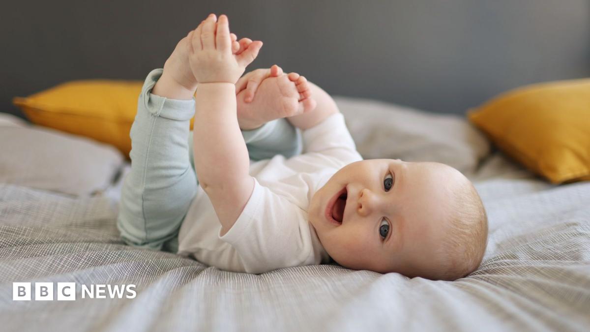 A baby is wearing a white top and blue leggings and is lying on a bed with yellow cushions.