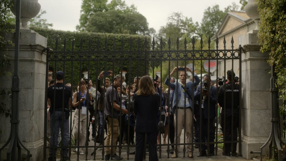 A crowd of reporters and security personnel gathers at a gated entrance, capturing the moment on their devices.