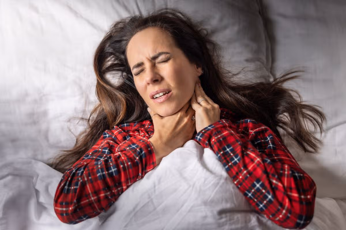 A woman in pain, with a pained expression on her face, holds her throat with both hands. The scene gives the impression that the person is in severe pain, probably due to illness or health problems.
