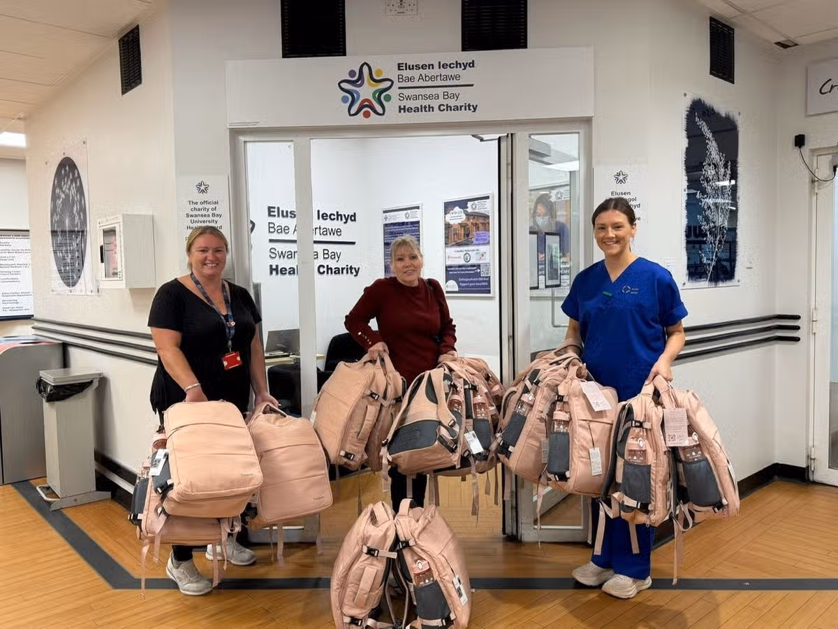 Three women standing in front of a doorway, holding pink rucksacks. There are 20 bags between them. The woman on the left is white. She is wearing a black top and navy lanyard, her blonde hair is tied back. The woman in the middle is white, in her fifties, her blonde hair also tied up. She has a dark red, long sleeved top on. The woman on the right is white with dark hair, tied back, she is wearing dark blue scrubs.
