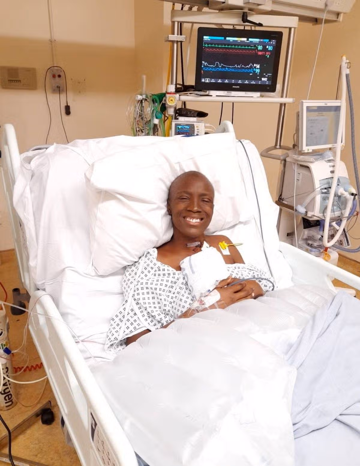 A person lies in a hospital bed, wearing a hospital gown and smiling while resting on pillows. The hospital bed is equipped with various medical devices, and a monitor is visible on a nearby stand.