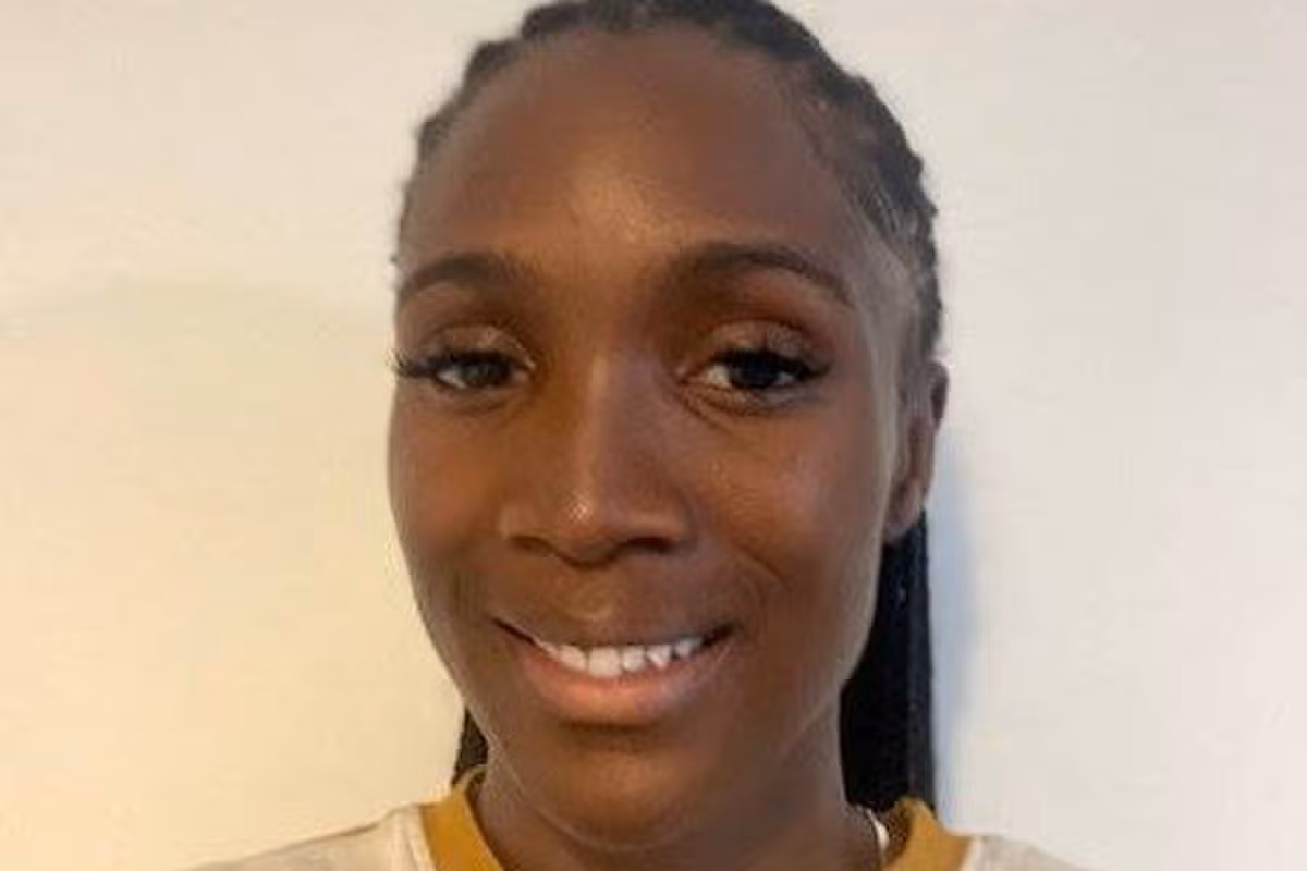 An individual stands before a plain background, wearing a multicolored shirt with a yellow accent. The person is smiling broadly, suggesting a cheerful demeanor. The individual's hair is styled in a way that it is partially visible over the shoulders.