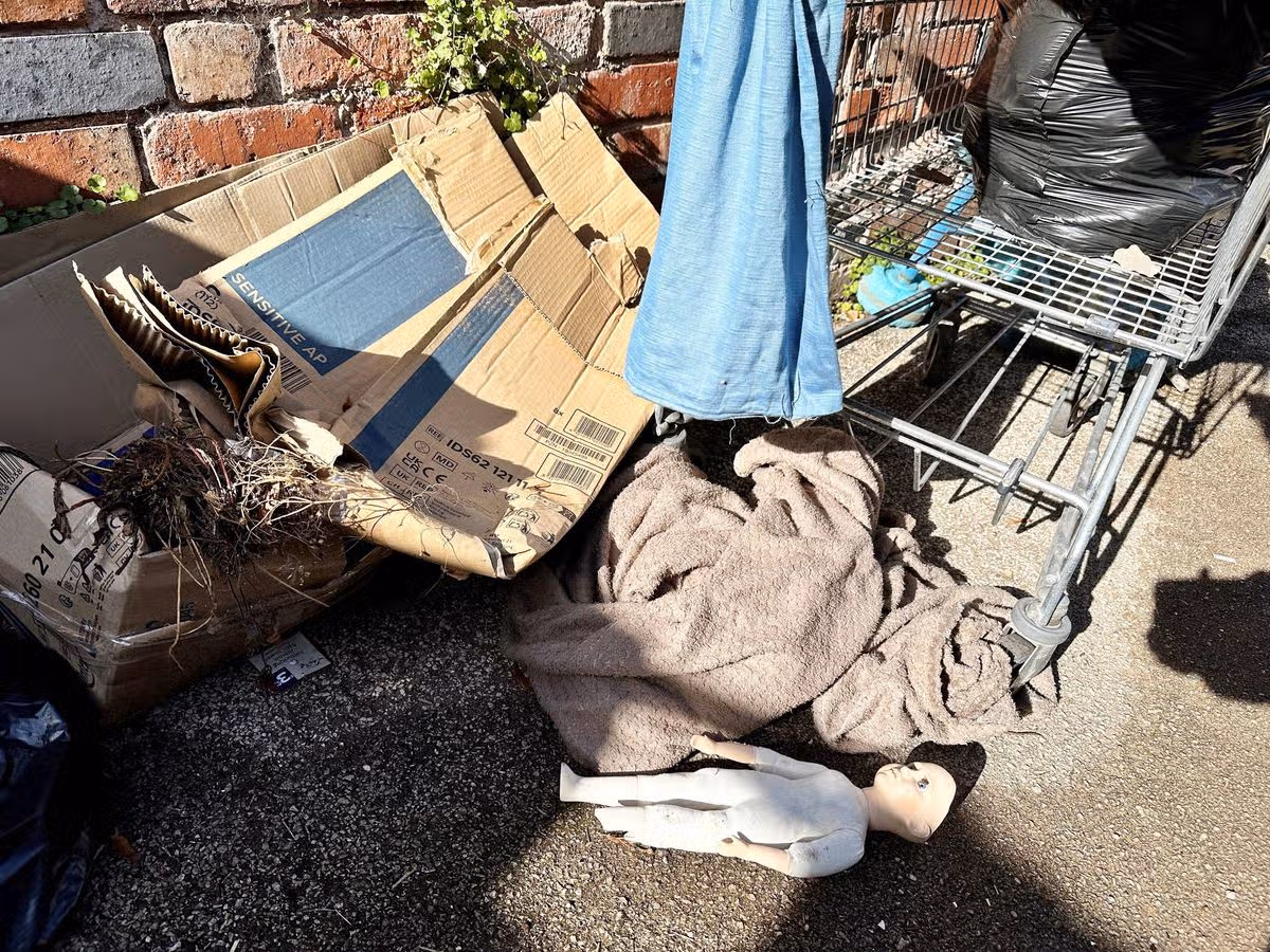 An assortment of discarded items, including a cardboard box, a blue cloth, and a plastic container, is scattered on the ground, adjacent to a brick wall.