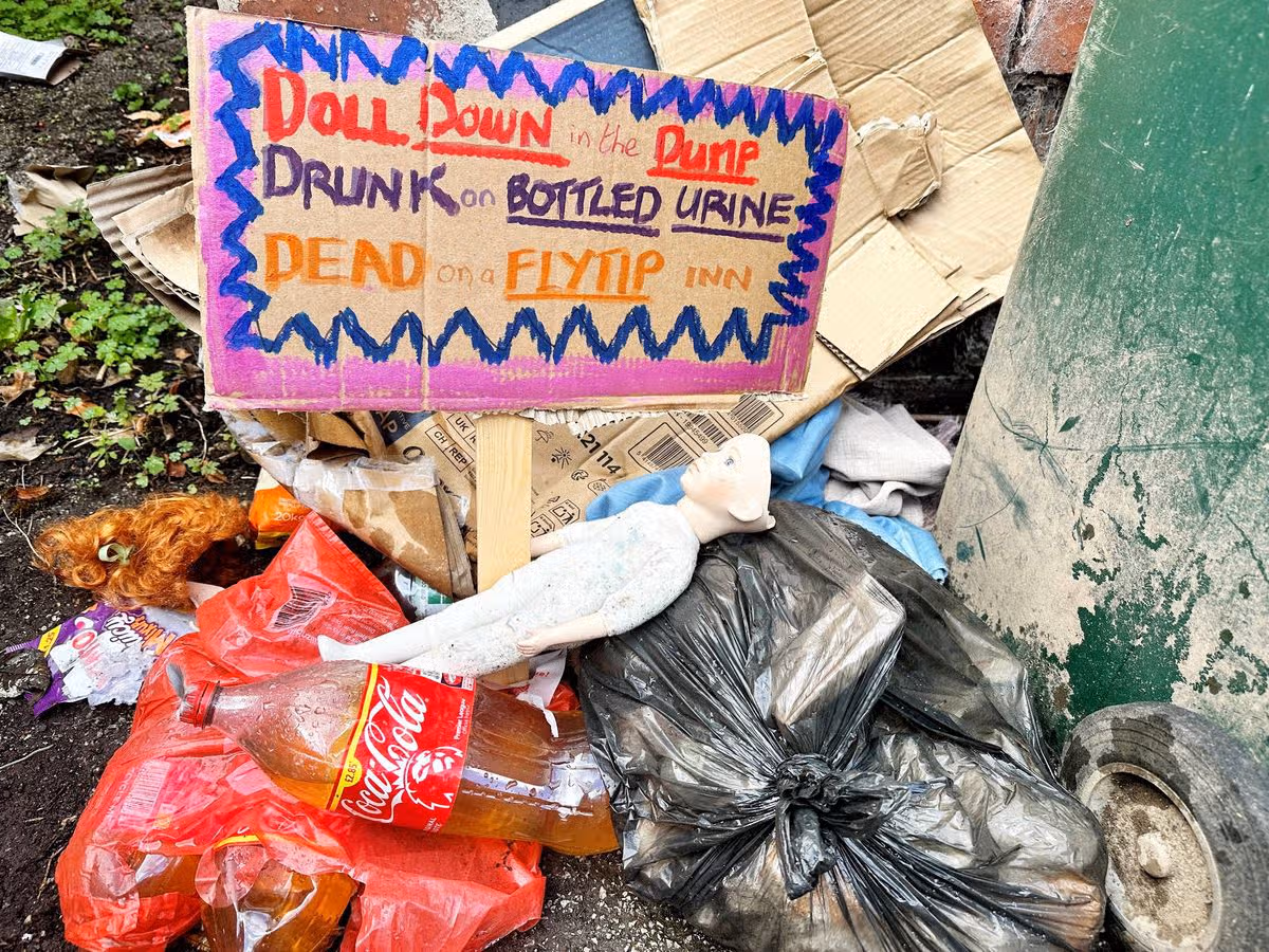 A pink sign with colorful lettering and a drawing of a bottle and a glass is placed amidst a collection of discarded materials, including plastic bags and other refuse, suggesting an area affected by littering and waste disposal issues.