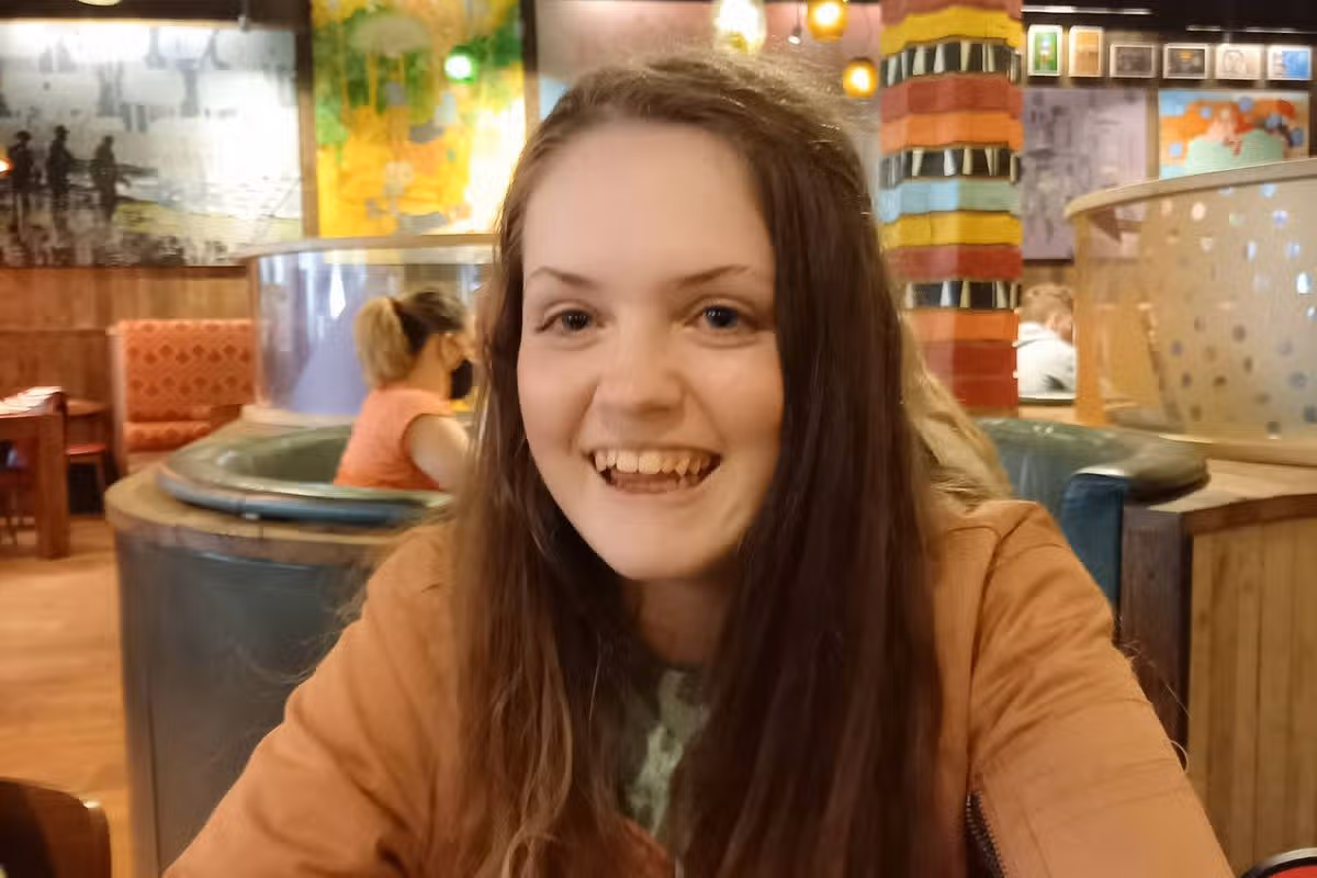 An individual with long dark hair and an orange top is seated in a restaurant, smiling and facing the camera.