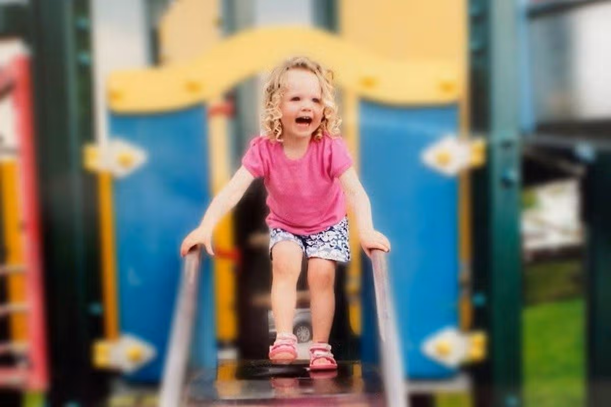 A child is playing on a slide in a playground, with a joyful expression on her face.
