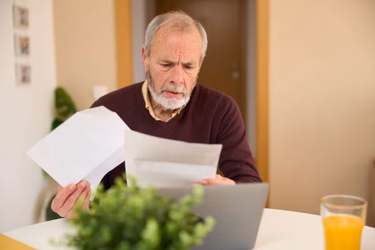 Focused senior man meticulously examining paperwork and using a laptop, managing finances or personal matters in a comfortable home setting