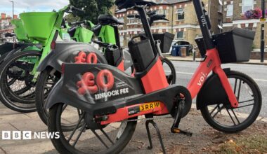 Image of a red Voi e-bike and green Lime bike parked on the side of a pavement. In the background there are rows of houses.