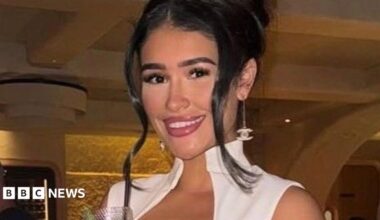 Alisha Samuels has long brown hair and is wearing ear rings and a white dress.