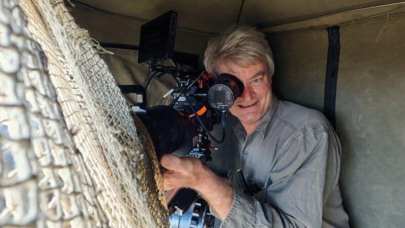 A man with gray hair looks into the viewfinder of a professional video camera while sitting inside a camouflaged blind.