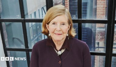 Lady Glenconner standing in front of a large black-framed window. She is looking up towards the camera smiling and is wearing a burgundy cardigan with a pearl necklace round her neck and a pair of pearl earrings.