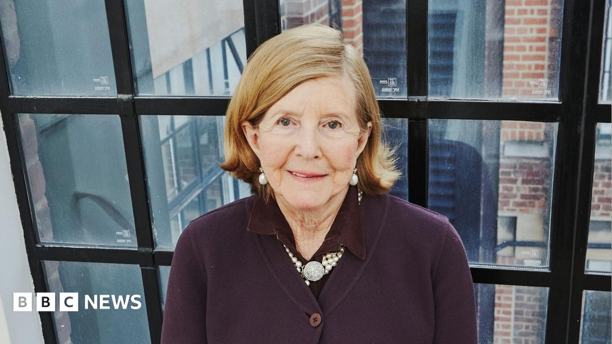 Lady Glenconner standing in front of a large black-framed window. She is looking up towards the camera smiling and is wearing a burgundy cardigan with a pearl necklace round her neck and a pair of pearl earrings.