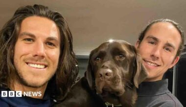 A young man with shoulder-length brown hair and light stubble smiling at the camera, and another young man to his right and a brown dog in between them