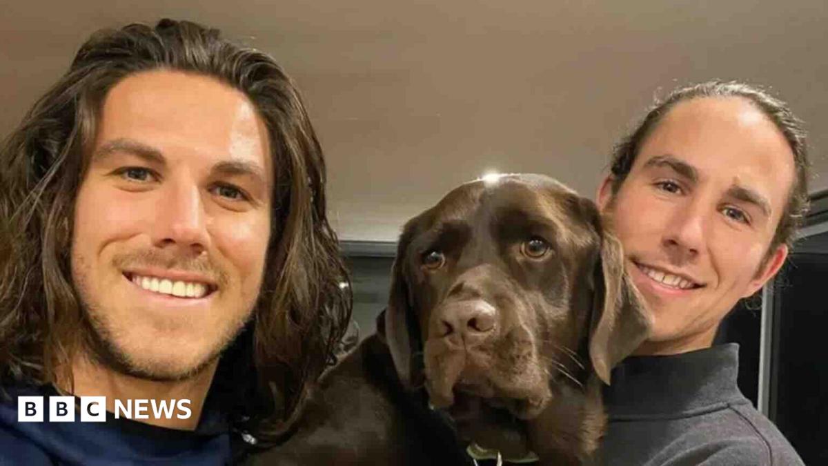 A young man with shoulder-length brown hair and light stubble smiling at the camera, and another young man to his right and a brown dog in between them