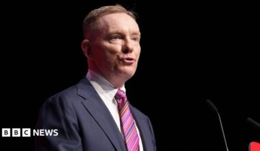 Sir Chris is talking into a mic on a stage. He's wearing a navy suit with a white shirt and stripy tie. The background is dark.