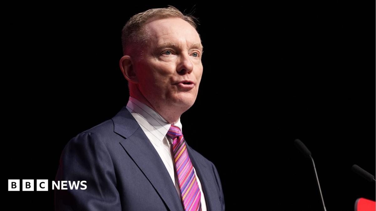 Sir Chris is talking into a mic on a stage. He's wearing a navy suit with a white shirt and stripy tie. The background is dark.