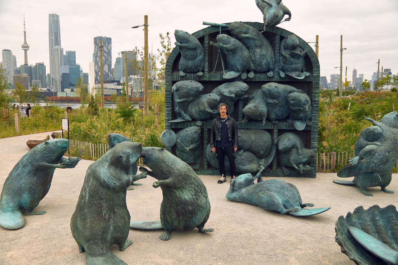 With the Toronto skyline in the rear, a large sculpture shows an array of beavers performing activities like dancing and playing drums. Some inhabit a large structure. A person with dark hair and a black leather jacket stands near the structure.