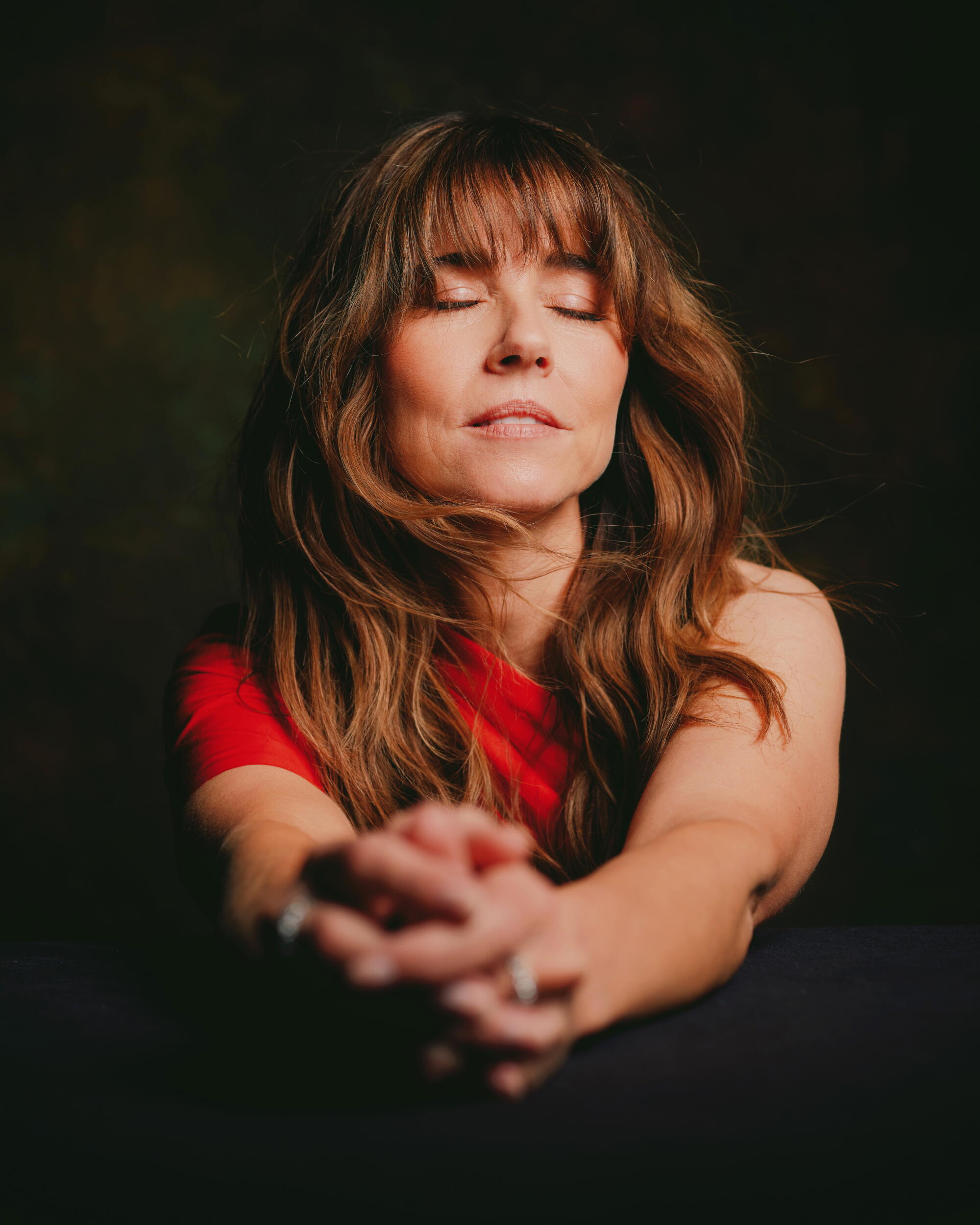 A woman with long flowing brown hair in a red dress with her eyes closed.