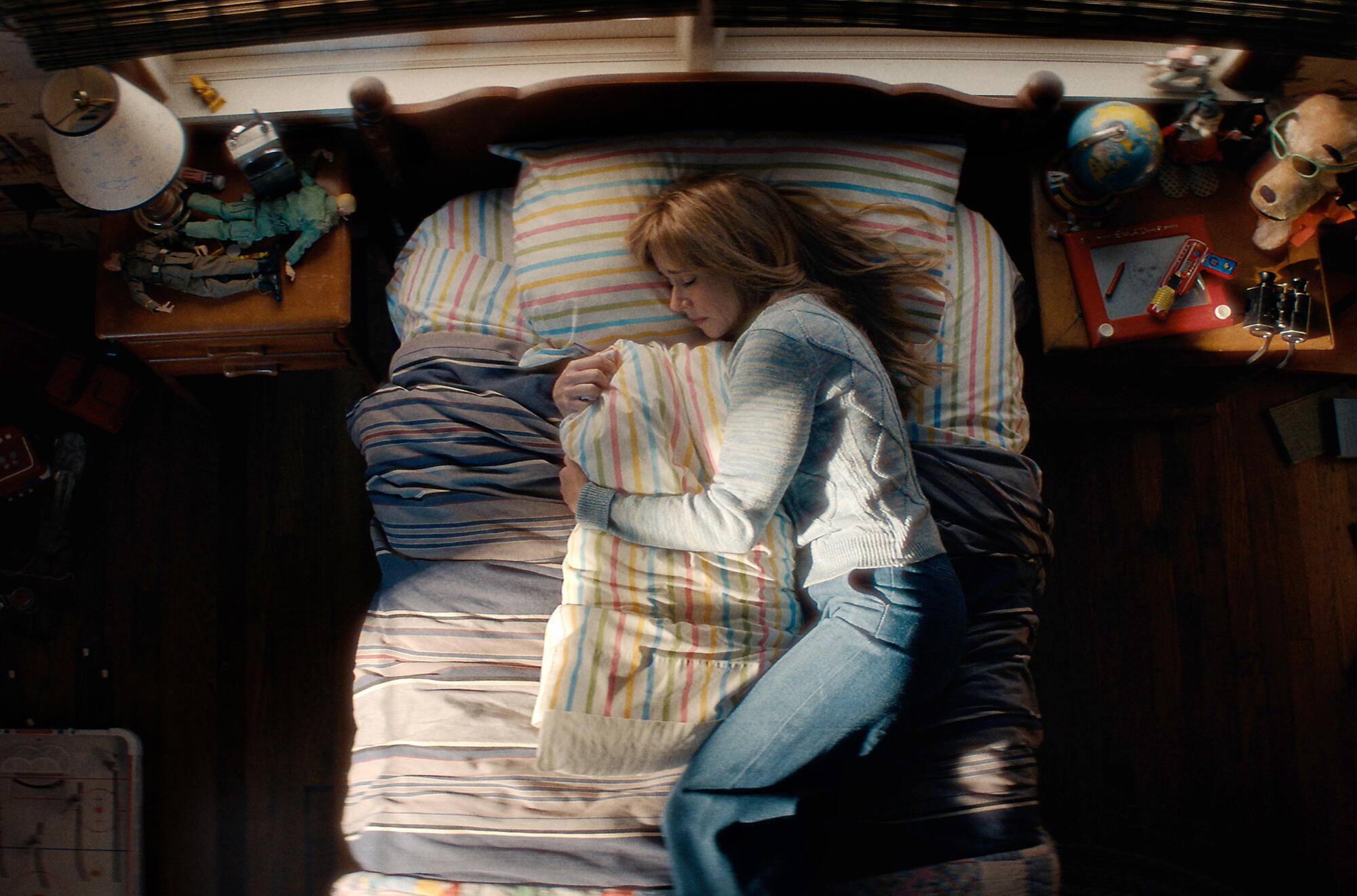 A woman in a blue sweater and jeans lays on a bed embracing a pillow.