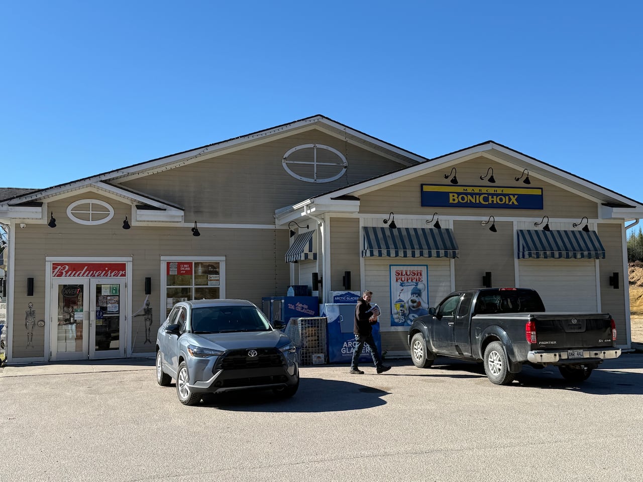 A beige building with Budweiser, Slush Puppie and Bonichoix signs with two cars parked out front while a man carrying items walks to his car.