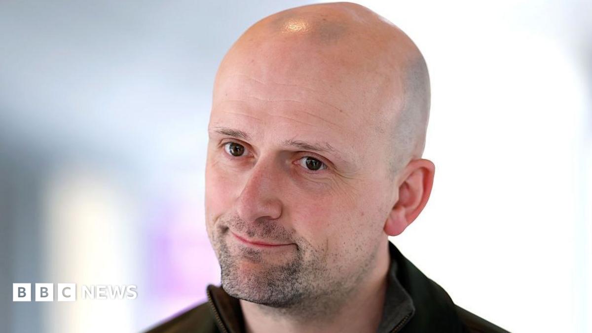 Stephen Flynn looking slightly off camera. Only his head and shoulders are visible and he appears to be wearing a dark top.