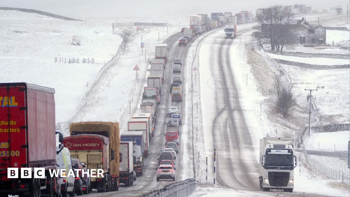 A road covered with snow and ice with snowy fields on either side, with a queue of cars and lorries in one direction and two lorries moving in the other.