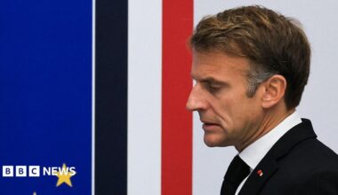 Close up of French President Emmanuel Macron taken from the side. He has short brown hair, and is wearing a white shirt, black jacket and tie.