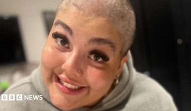 A woman with a shaved head and long eyelashes smiles at the camera. She is wearing a grey hoodie.