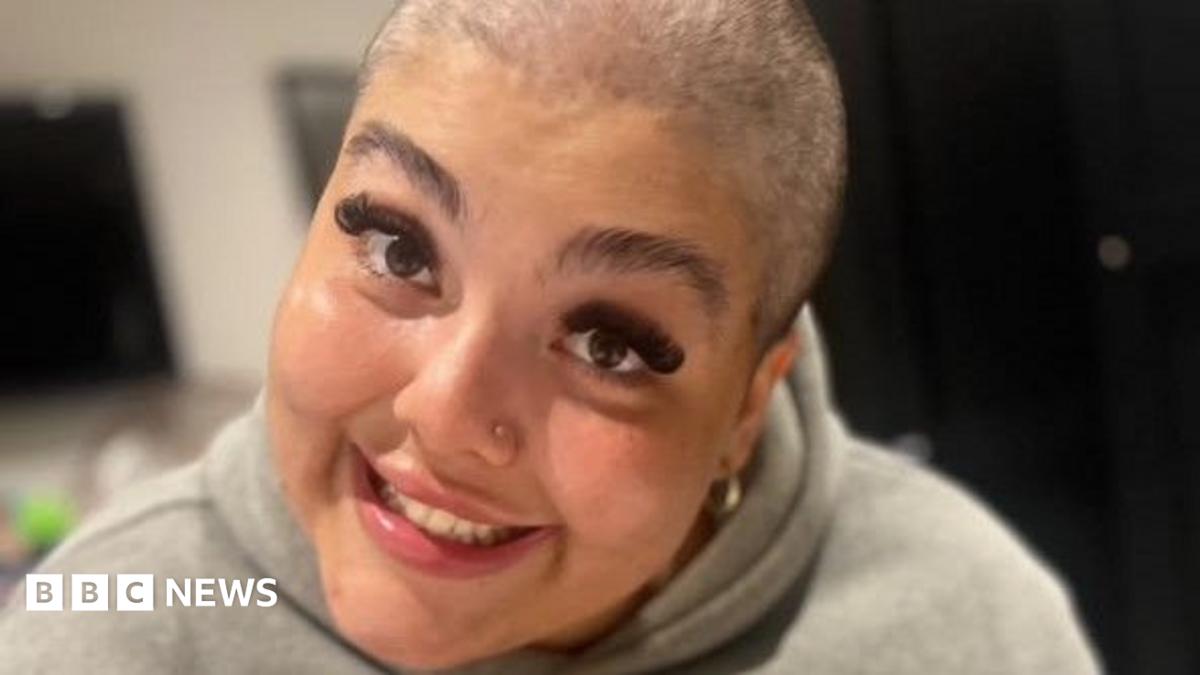 A woman with a shaved head and long eyelashes smiles at the camera. She is wearing a grey hoodie.