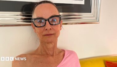 A woman in her 50s is sat on a yellow sofa with orange pillows. She is wearing a bright pink, off the shoulder, long-sleeved top, and glasses with thick black frames. Her hair is tied up, and she's looking at the camera with a faint smile. Behind her you can see the bottom of a large silver framed photo.