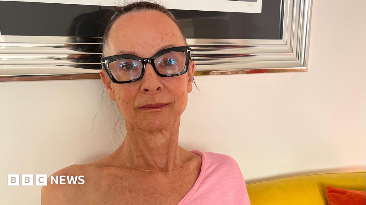 A woman in her 50s is sat on a yellow sofa with orange pillows. She is wearing a bright pink, off the shoulder, long-sleeved top, and glasses with thick black frames. Her hair is tied up, and she's looking at the camera with a faint smile. Behind her you can see the bottom of a large silver framed photo.